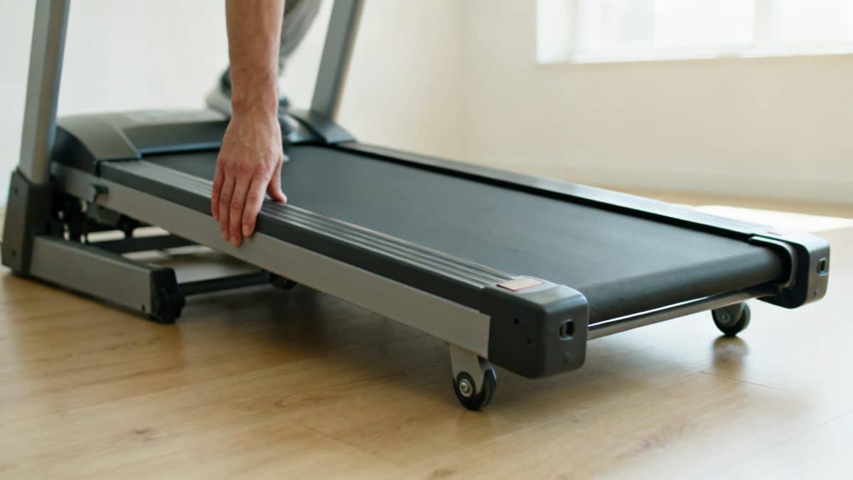Un tapis de course incliné vu de profil, une main posée sur la rampe latérale.